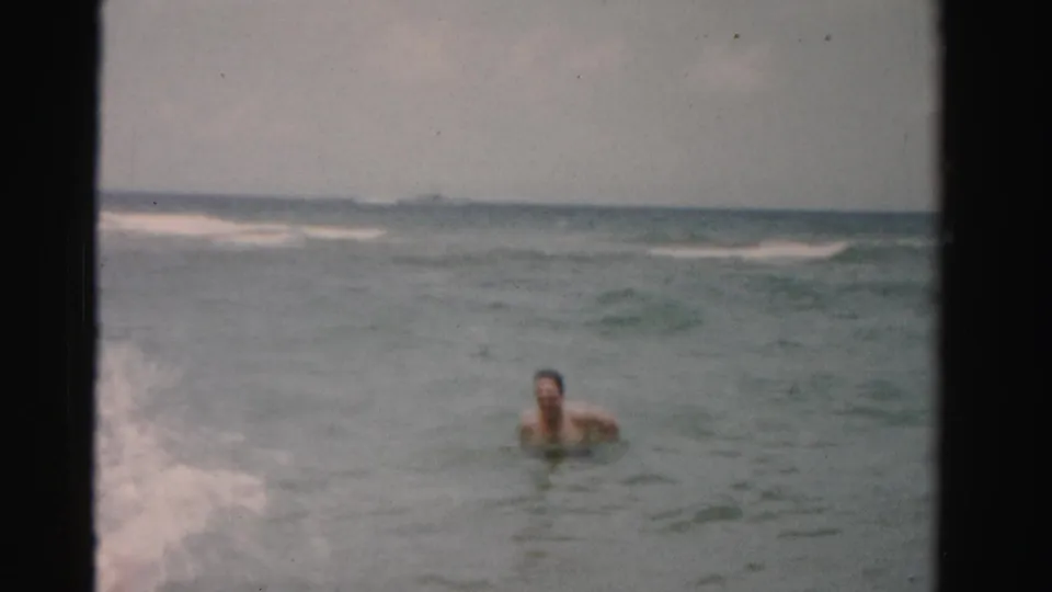 1955 8mm frame of an adult man swimming near a breaking wave off Dade County, Florida, seen from a handheld shoreline viewpoint.