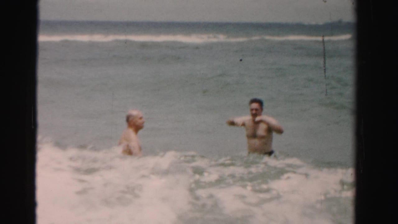 1955 Dade County, Florida 8mm frame showing two adult men standing chest-deep and waist-deep in choppy ocean surf.