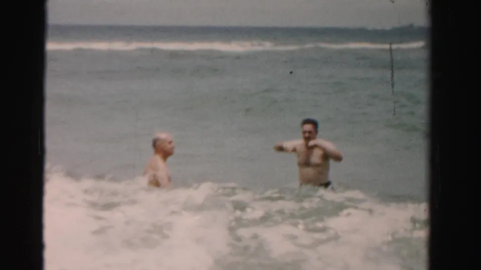 1955 Dade County, Florida 8mm frame showing two adult men standing chest-deep and waist-deep in choppy ocean surf.