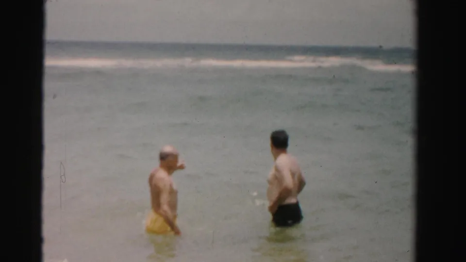 1955 Dade County, Florida home-movie frame of two adult men facing each other in the ocean, one gesturing toward the water.