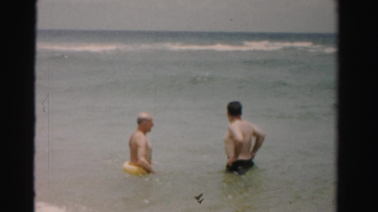Two adult men stand in shallow ocean water, one with a yellow swim ring, with waves behind them.