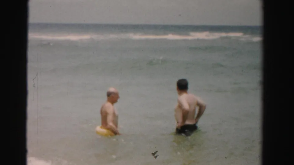In 1955 Dade County, Florida, two adult men stand in shallow surf, one wearing or holding a yellow swim ring as small waves break offshore.
