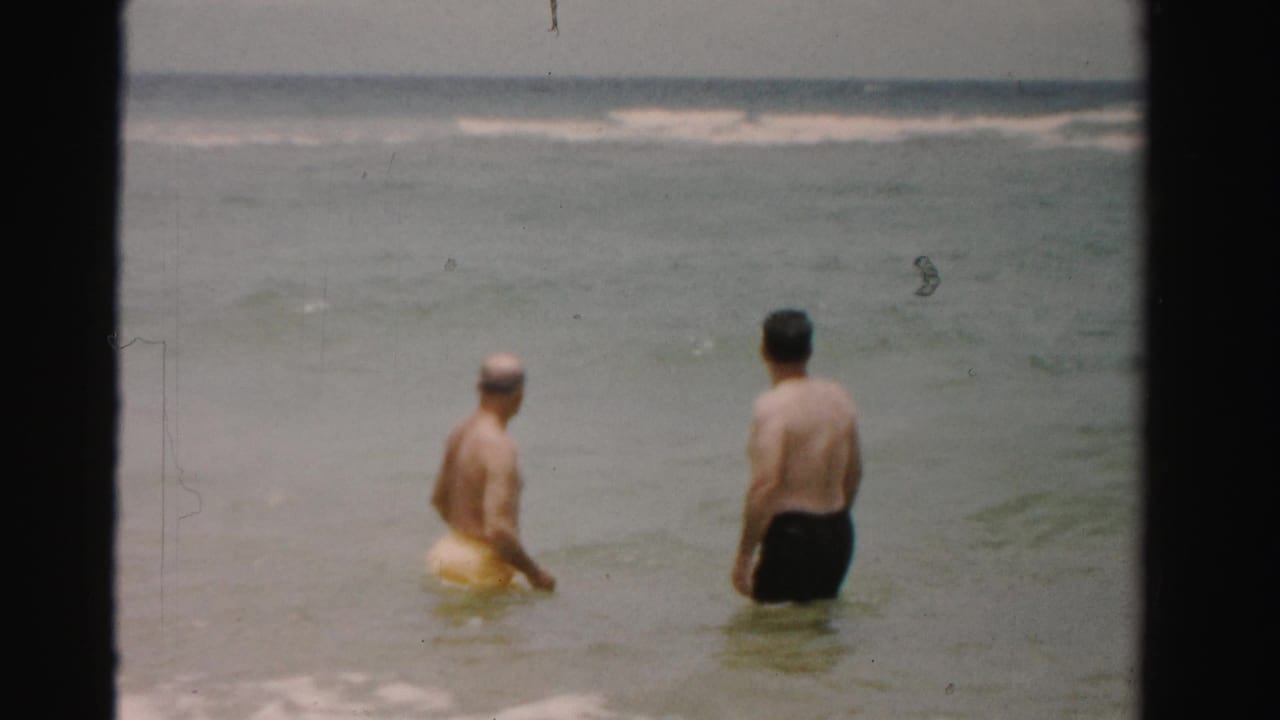 Two adult men face the water in shallow surf, with one man beside a yellow swim ring.