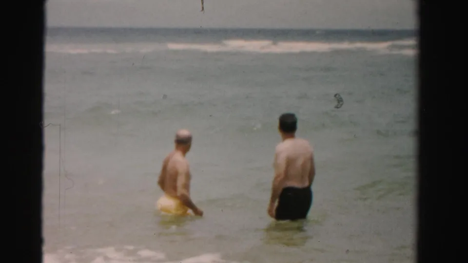 Two adult men stand waist-deep in the ocean in Dade County, Florida, with one beside a yellow swim ring and small breakers in the distance.