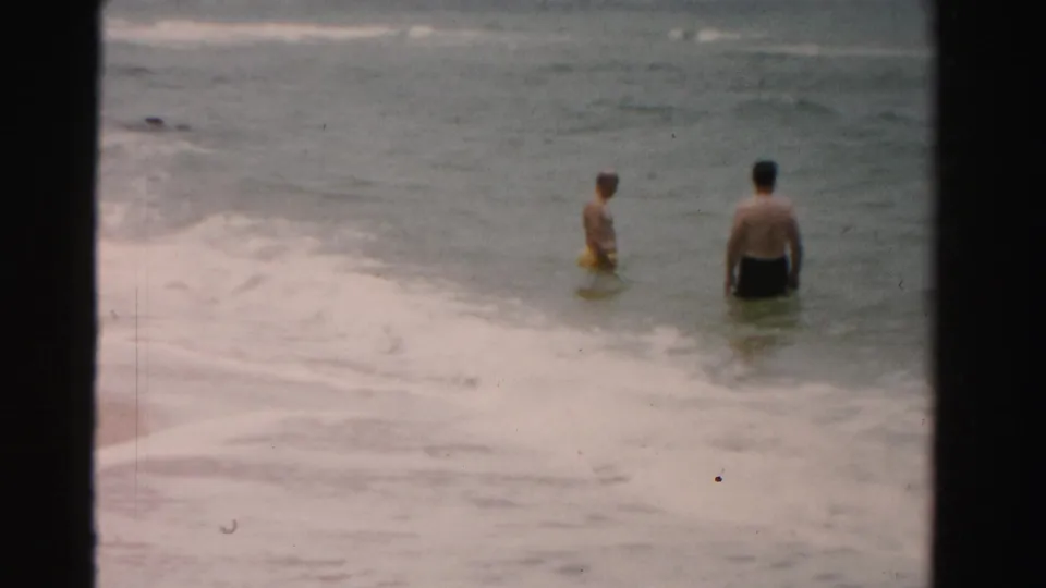 From a slightly wider 8mm view, two adult men stand in the Dade County surf while foamy water rolls across the sandy shoreline.