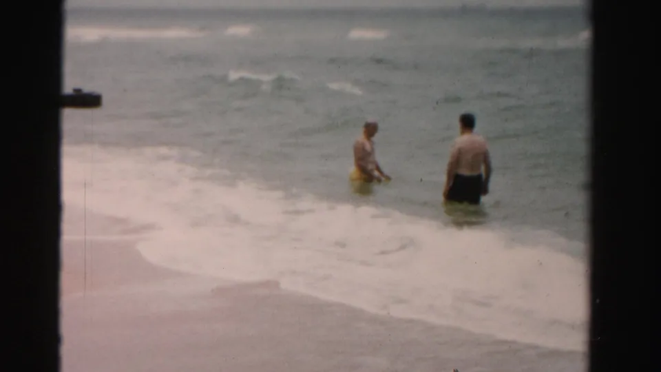 Two adult men stand near the foamy shoreline in 1955 Dade County beach footage, one with a yellow swim ring as waves break behind them.