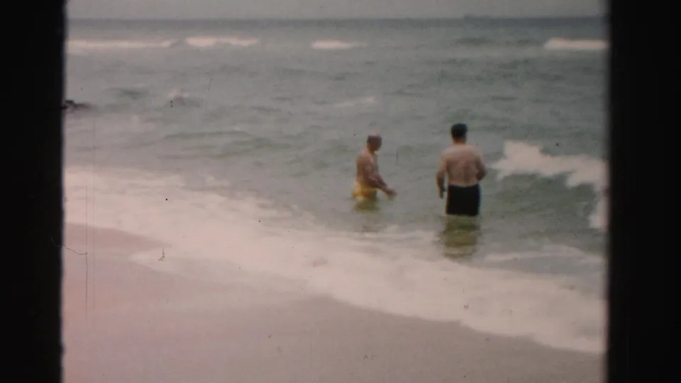 Two men stand in shallow ocean waves at a 1955 Dade County, Florida beach, framed by dark 8mm film edges.