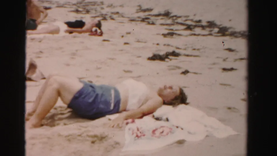 A woman in a white top and blue shorts sunbathes on a sandy Dade County beach with towels and seaweed nearby.