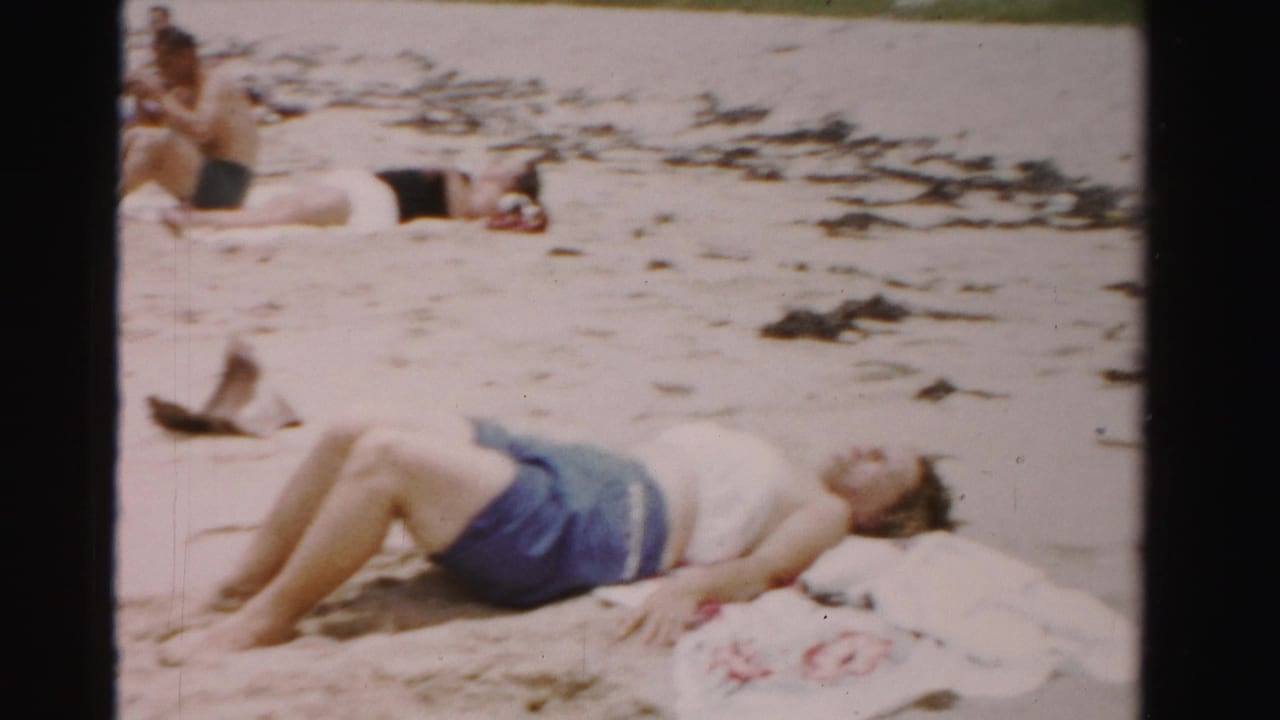 Woman lying on a beach towel in the sand with other beachgoers behind her.