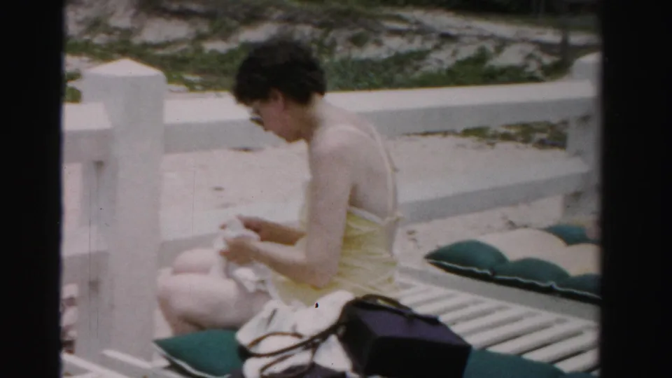 In this 1955 Dade County, Florida beach frame, a woman in a yellow swimsuit sits on a white lounge chair with a towel and bag nearby.
