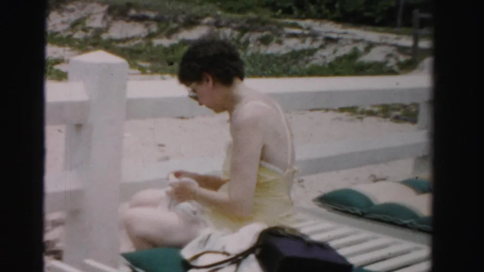 A 1955 Dade County, Florida home-movie frame shows a woman seated on a white beach lounge chair, looking down at a towel.