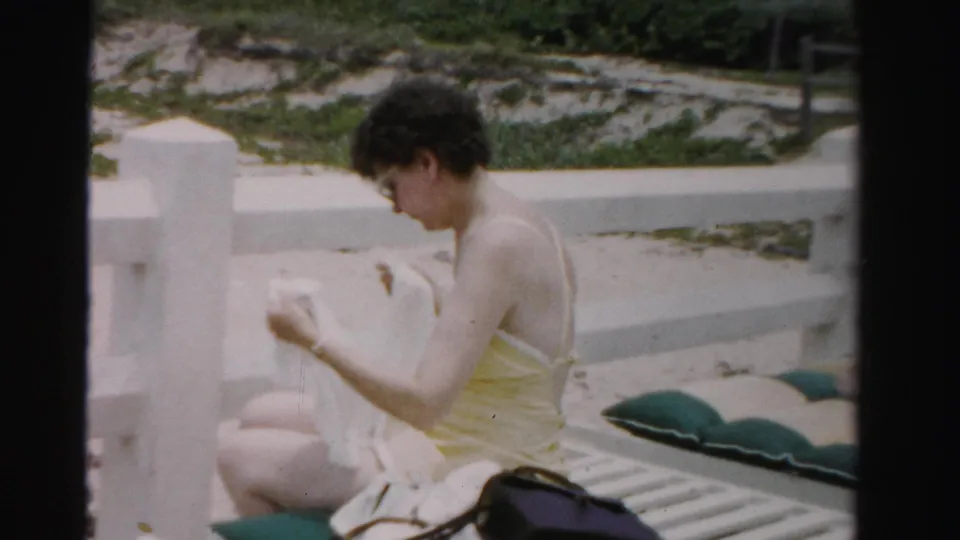 In this 1955 Dade County, Florida beach scene, a woman on a white lounge chair raises a towel while seated beside green cushions.