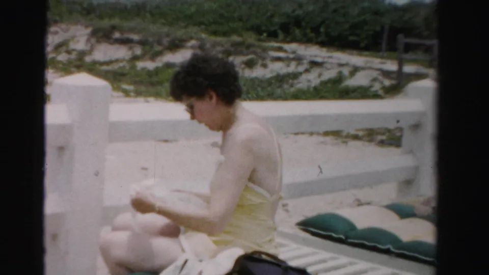 1955 Dade County beach home-movie frame showing a woman in a yellow swimsuit seated on a striped lounge chair near cushions and a low wall.