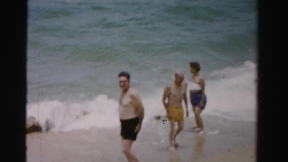 1955 Dade County beach frame showing two men and a woman walking through shallow water along the surf line.
