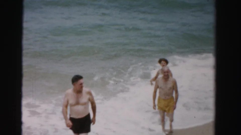 1955 Dade County home-movie frame with two men walking through the foamy shoreline while a woman follows near the waves.