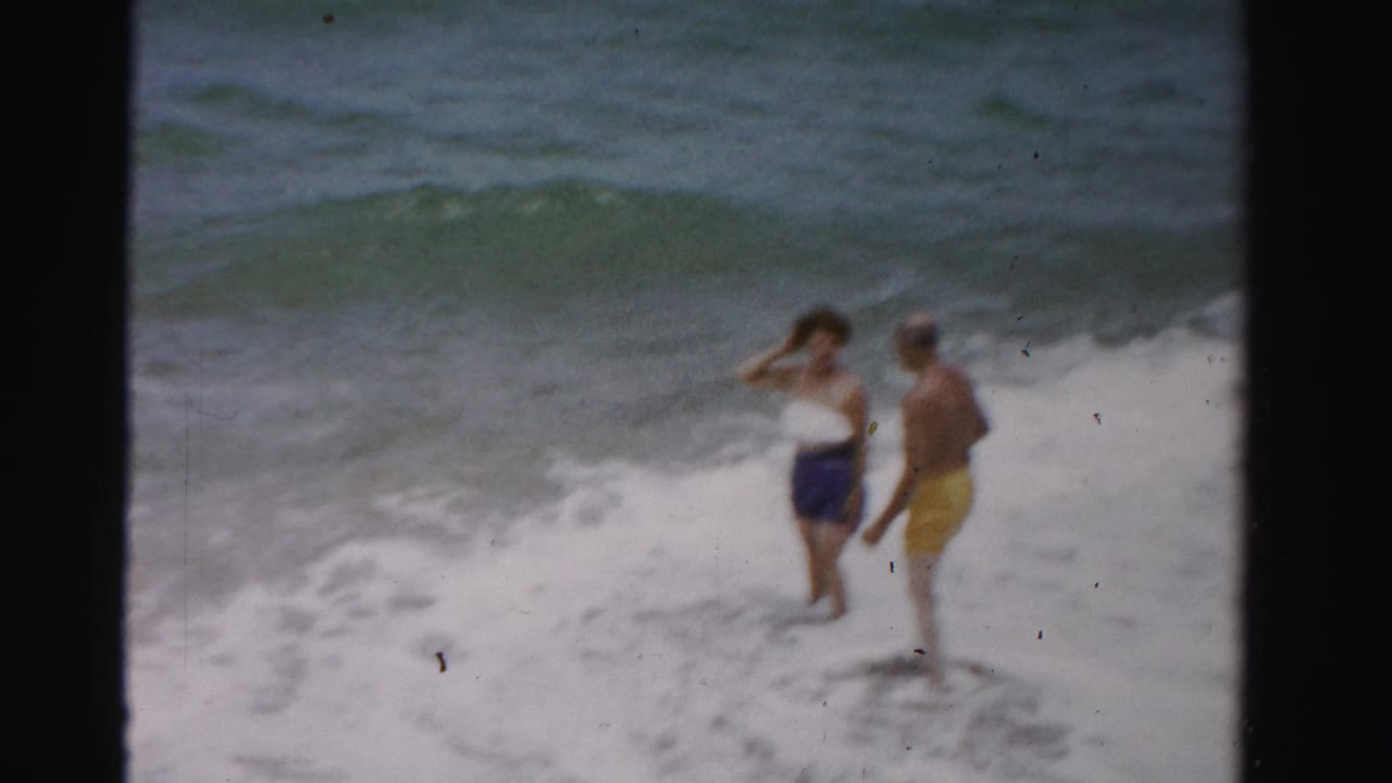 Two adults stand in shallow ocean surf with waves behind them.
