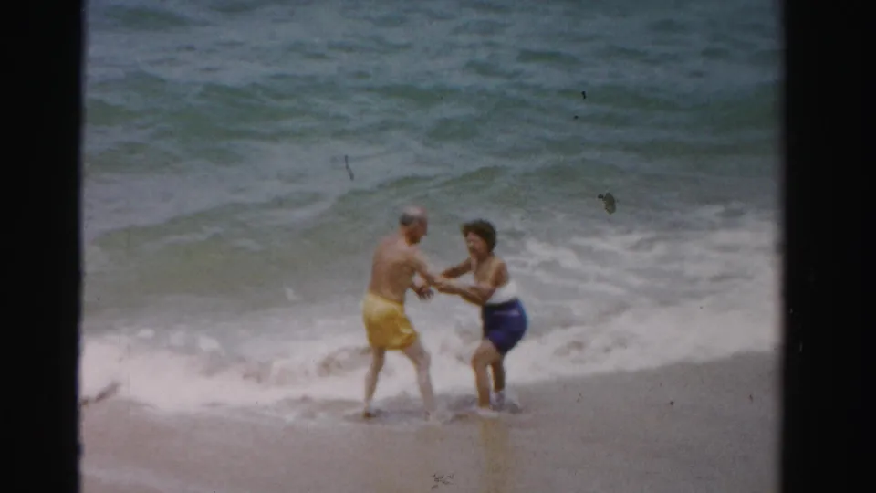 The couple holds hands in shallow Dade County surf, with 8mm grain and faded color visible across the frame.
