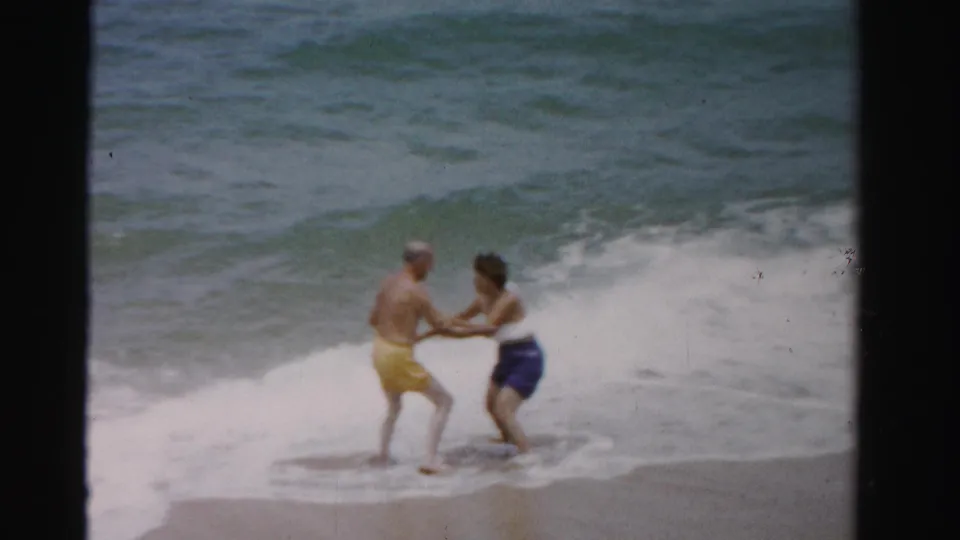 A man and woman hold hands at the shoreline as a wave washes in, captured in 1955 Florida 8mm home-movie footage.