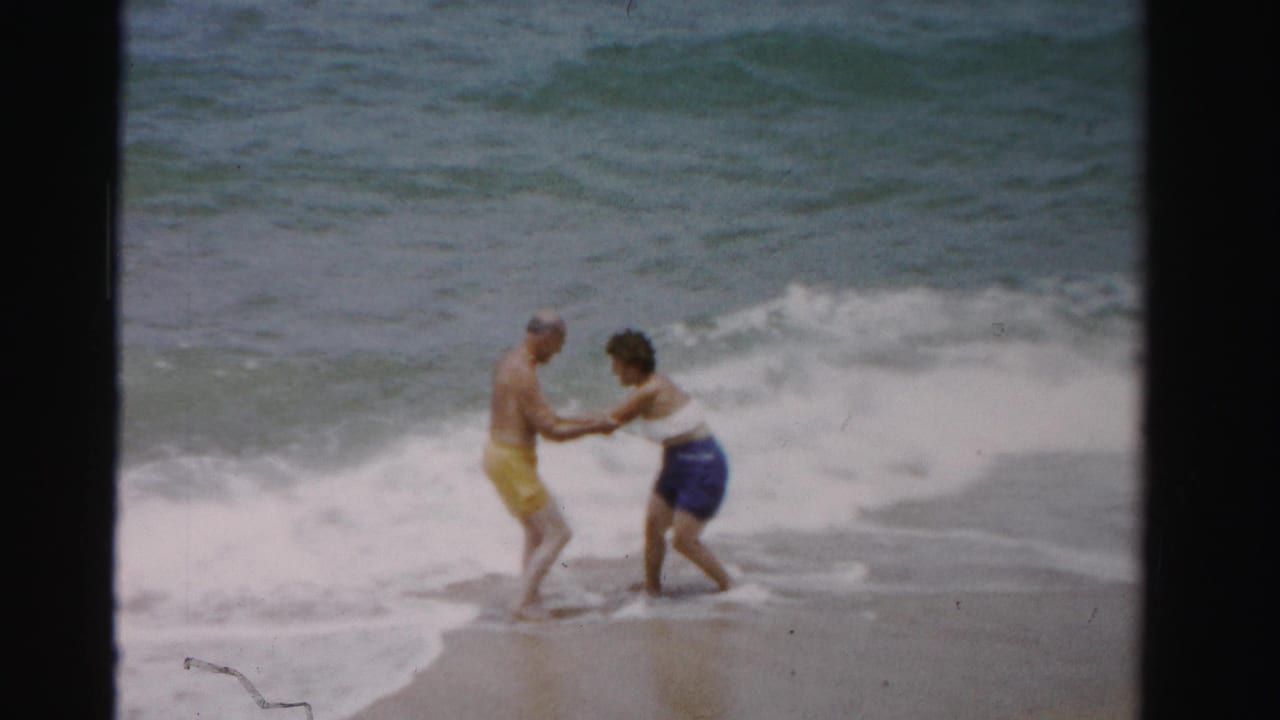 Two adults hold hands in shallow ocean surf near wet sand.