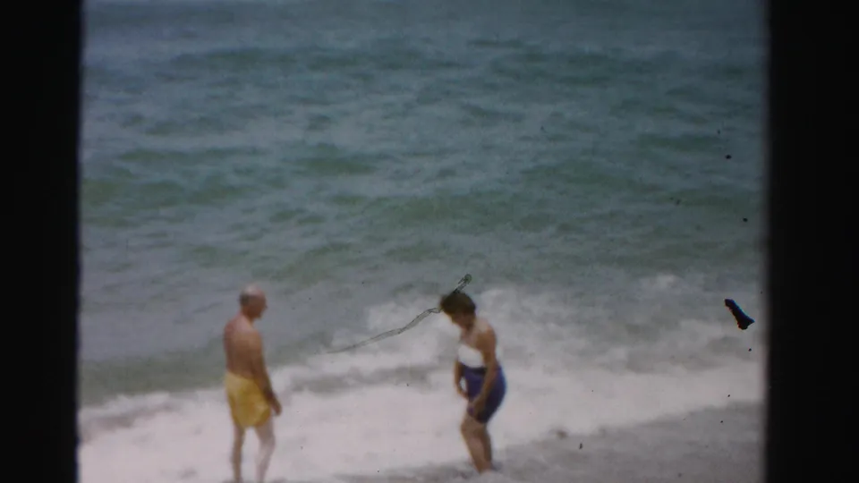 The man in yellow trunks and woman in blue shorts stand in the shallow Dade County surf during a playful pause.
