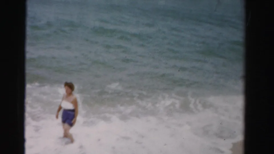 A woman in a white top and blue shorts stands alone in shallow surf in the 1955 Dade County beach scene.