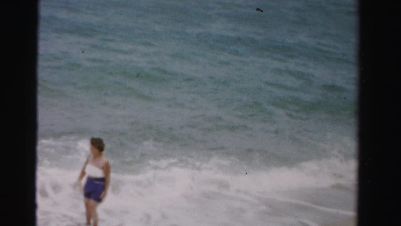 Woman in a white top and blue shorts walking near ocean surf.