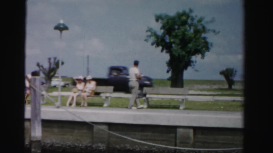 A Dade County waterfront walkway shows visitors on benches, a man walking past, trimmed trees, and a dark mid-century car near the water.