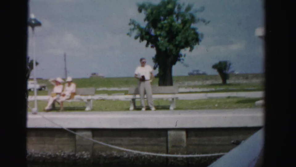 1955 Dade County waterfront park scene with seated visitors, a standing man, benches, a tree, and harbor water beyond.