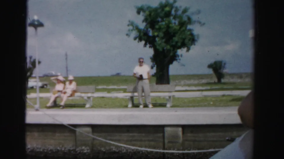 Miami-area waterfront park frame with a standing man, seated visitors, benches, a tree, and cloudy sky in 1955 home-movie footage.
