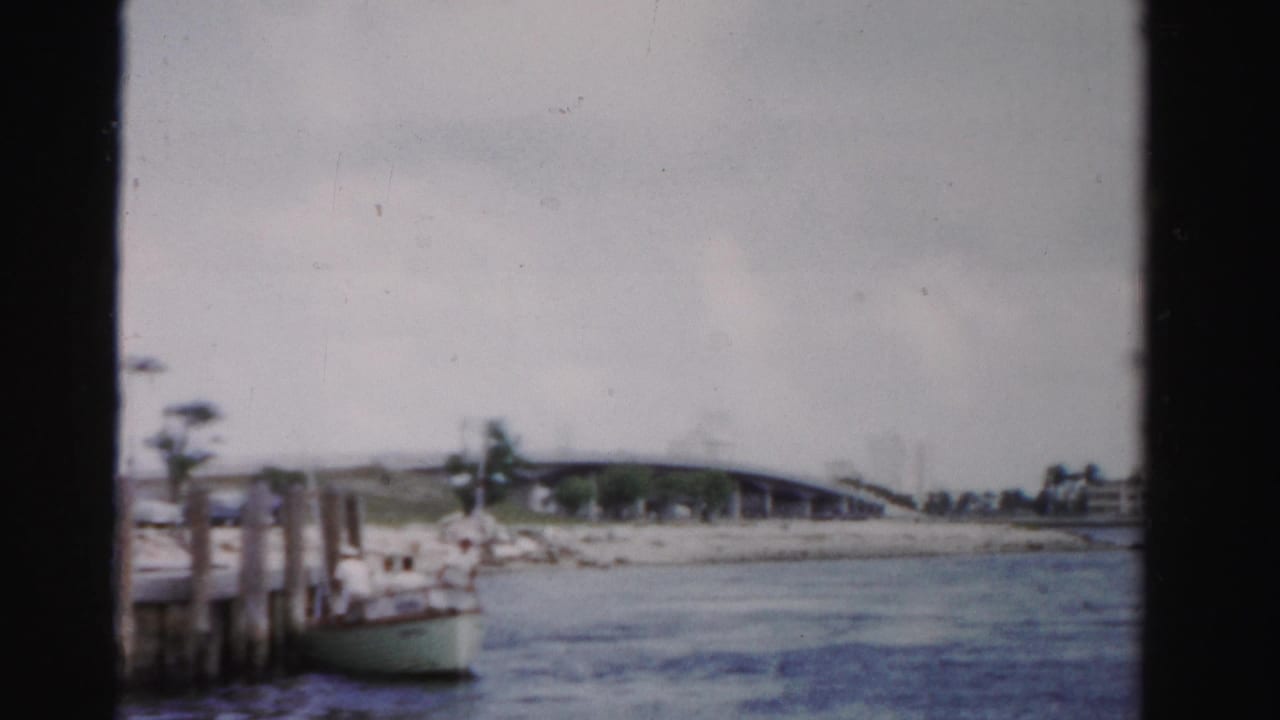 Canal view from a boat with a low bridge, shoreline, dock posts, and a small moored boat.