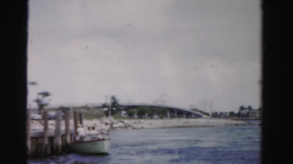 1955 Dade County canal scene from a moving boat, showing water, a low bridge, shoreline palms, dock posts, and a small boat.