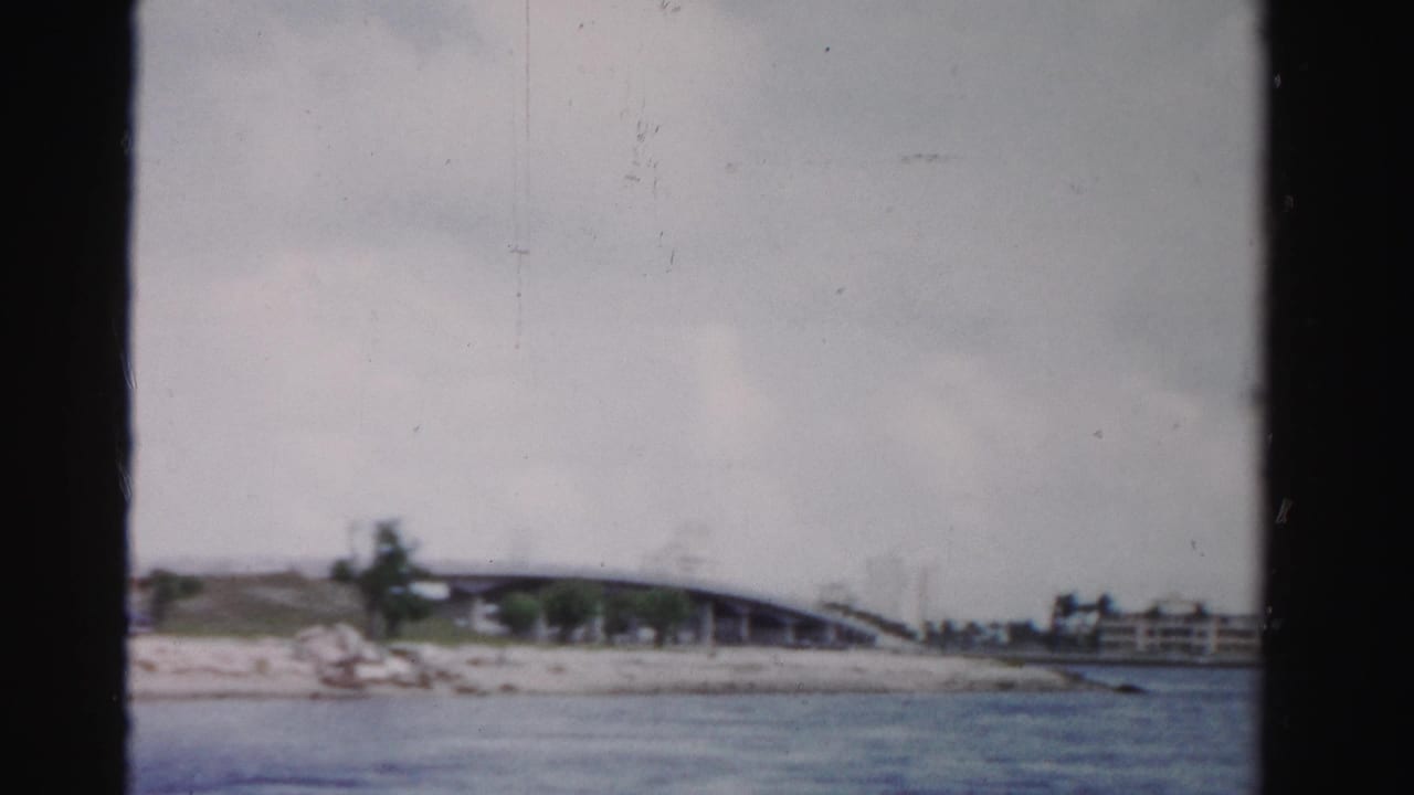 Canal water with a low bridge, shoreline vegetation, and distant waterfront buildings.