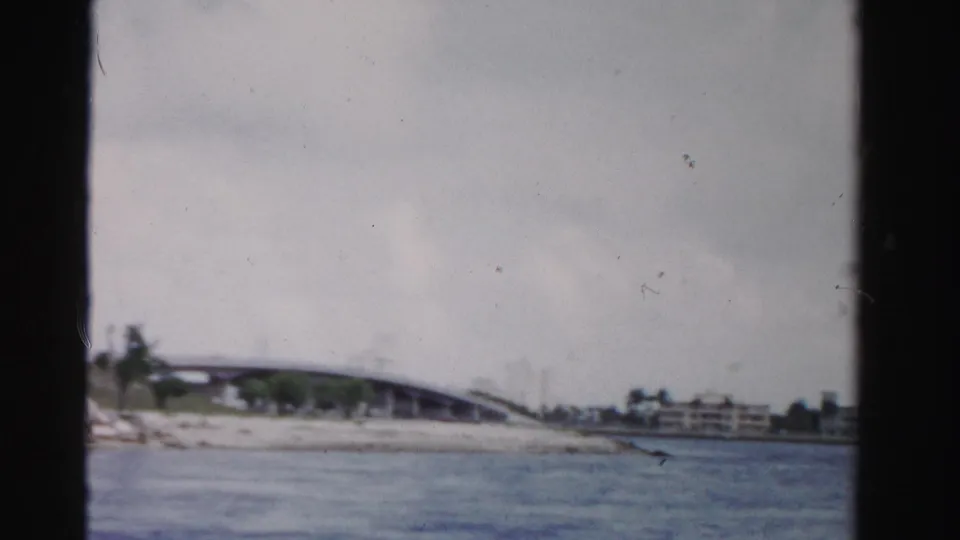 Soft 8mm frame of a Dade County canal in 1955, with a bridge, open water, low shoreline, and distant buildings.