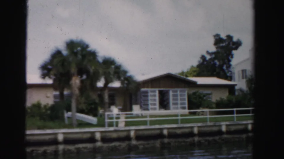 1955 Dade County waterfront home seen from a boat, with palm trees, a white railing, dock edge, and canal water.