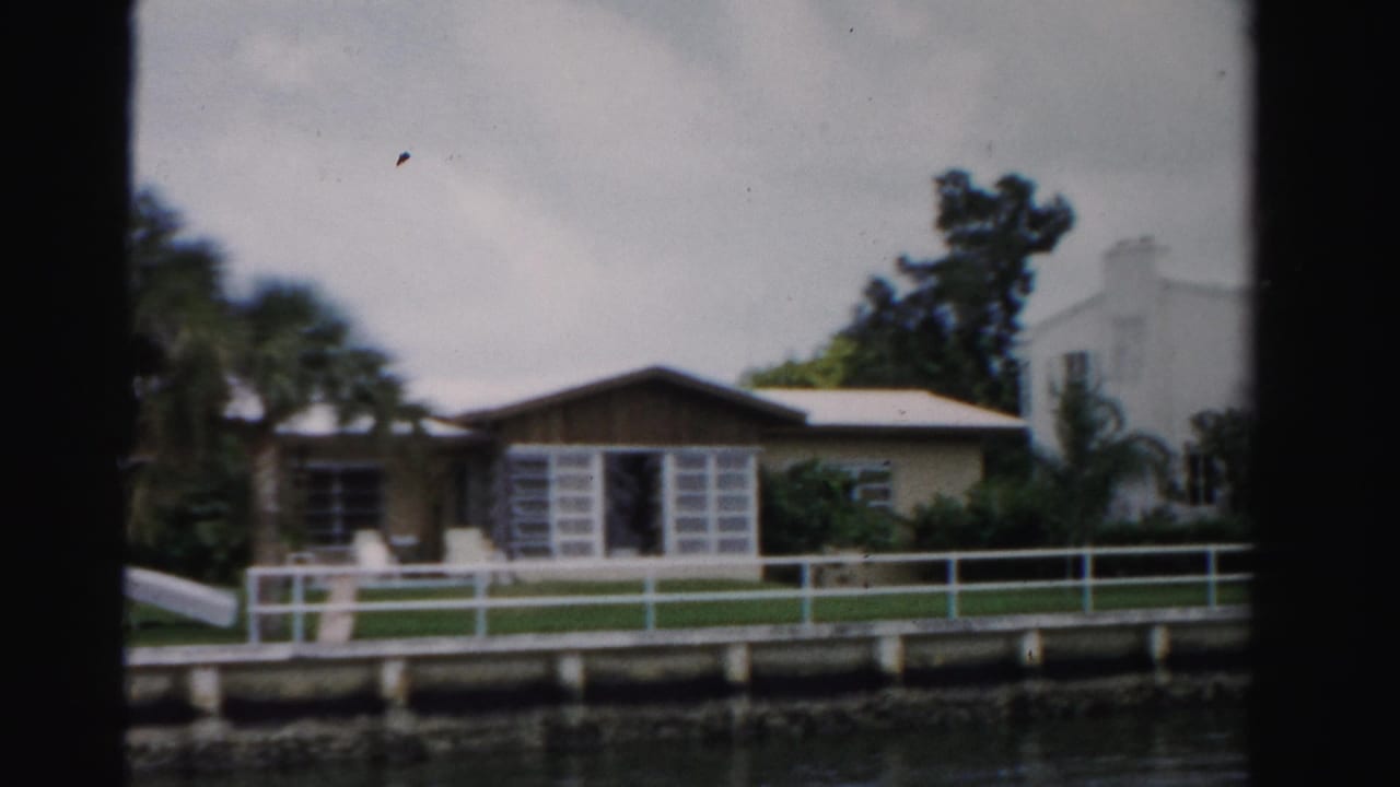 Waterfront house with white railing, canal edge, lawn, trees, and cloudy sky.