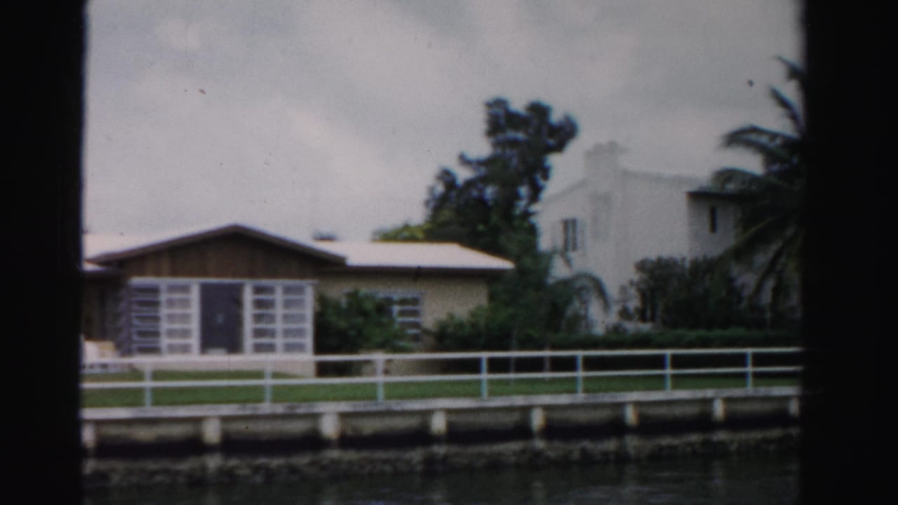 Canal-side house with white railing, hedge, trees, and pale building behind it.