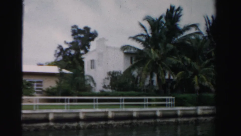 1955 Dade County waterfront frame with a white building partly hidden by palms, hedges, railing, canal edge, and cloudy sky.