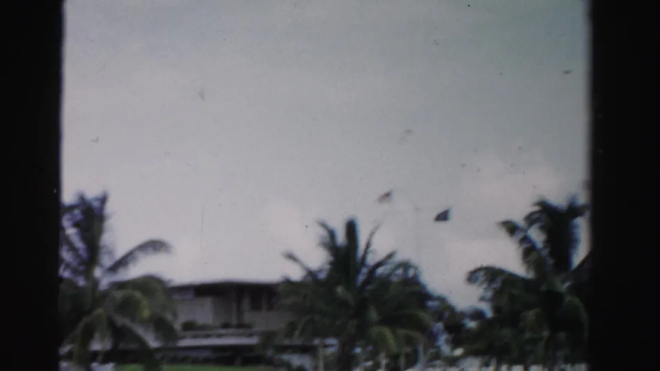 1955 Dade County home-movie frame showing a low waterfront house behind palm trees under a cloudy sky.