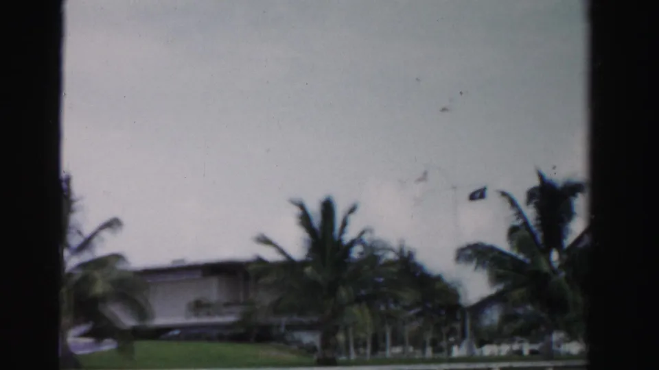 1955 Dade County frame with a modern low house, palms, lawn, and overcast sky viewed from a fixed home-movie angle.