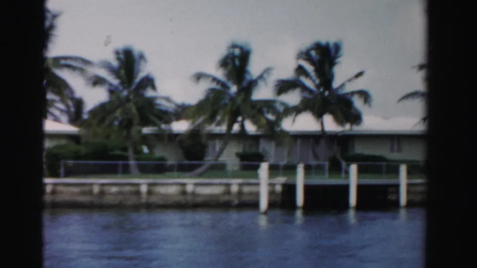1955 Dade County waterfront view showing a low house, palm trees, dock posts, seawall, and calm canal or lake water.