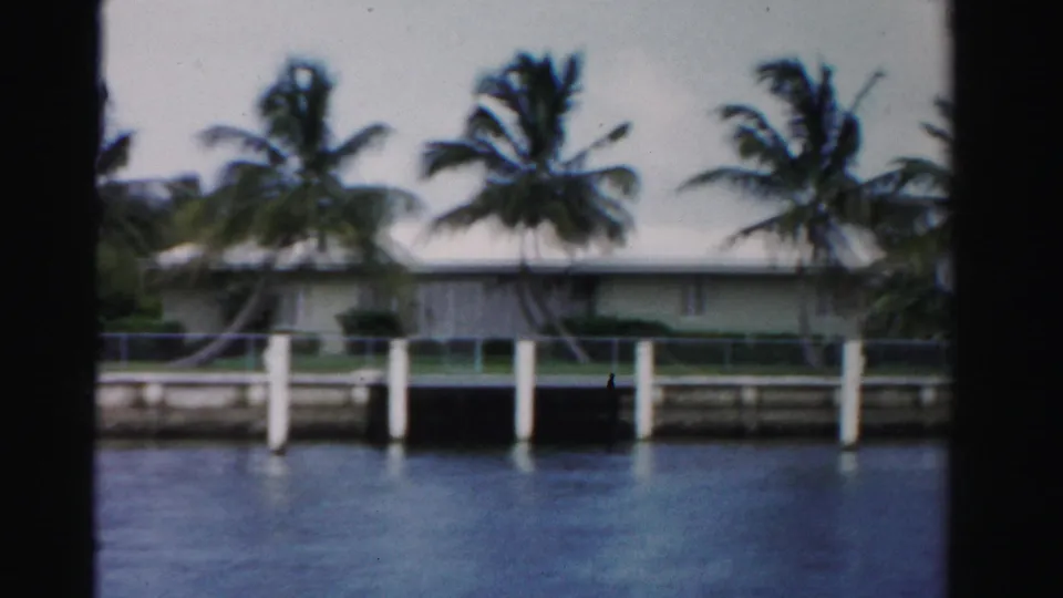 1955 Dade County home-movie frame of a palm-lined waterfront house with white dock posts and a seawall across blue water.