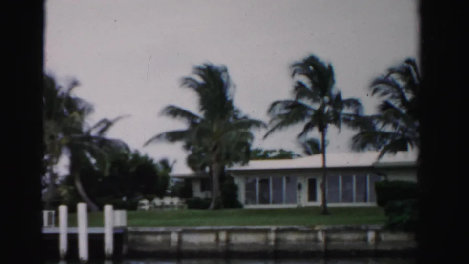 1955 Dade County home-movie frame showing a canal-side lawn, dock pilings, palms in wind, and a low residence with large windows.
