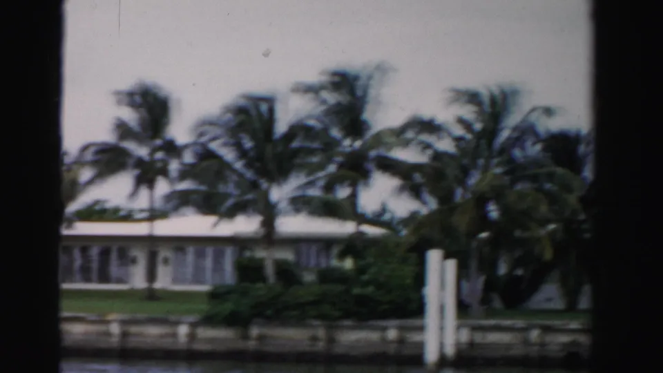 1955 Dade County frame with a low waterfront home, palms, shrubs, seawall, and white dock pilings viewed through dark 8mm frame edges.