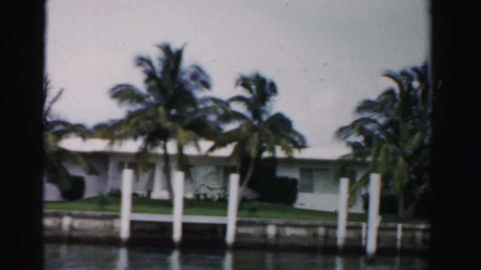 Dade County canal-side home with white pilings, palm trees, lawn, and a low mid-century roofline in soft 8mm focus.