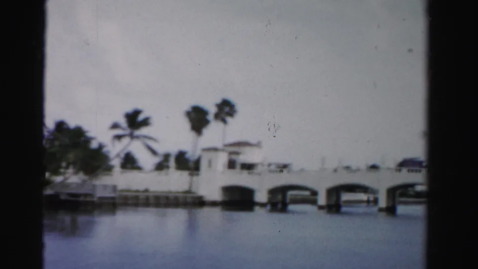 White arched bridge spanning a canal in Dade County, with palms and low waterfront structures visible behind it.