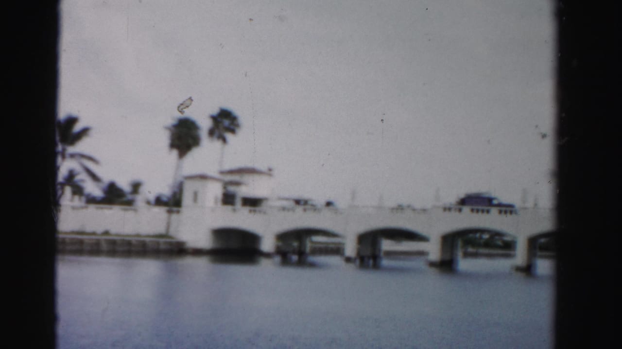 1955 Dade County 8mm frame showing a white arched bridge over water, palm trees behind it, and a dark car crossing.