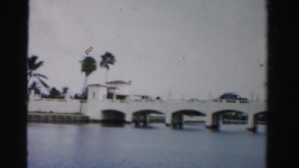 1955 Dade County 8mm frame showing a white arched bridge over water, palm trees behind it, and a dark car crossing.