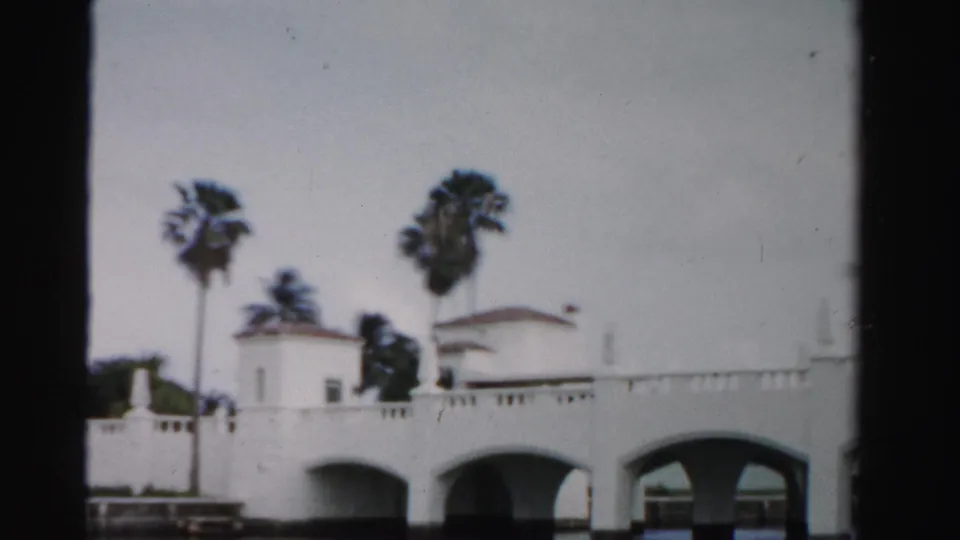 1955 Dade County 8mm frame with a white arched bridge, palm trees, calm water, and small bridge-side structures.