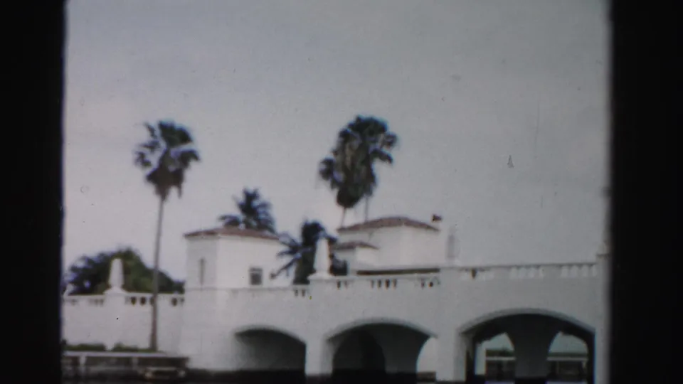 1955 Dade County 8mm frame showing a white arched bridge, pale stucco buildings, and palms viewed from across the water.