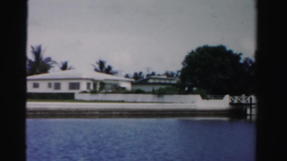 1955 Dade County waterfront frame with a low white house, dense trees, a small dock or landing, and rippled canal water.